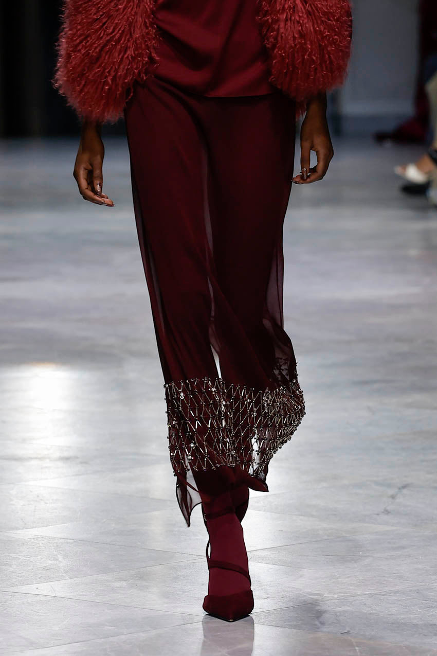 Model walking on a runway wearing burgundy pants with lace hem and red fur coat.