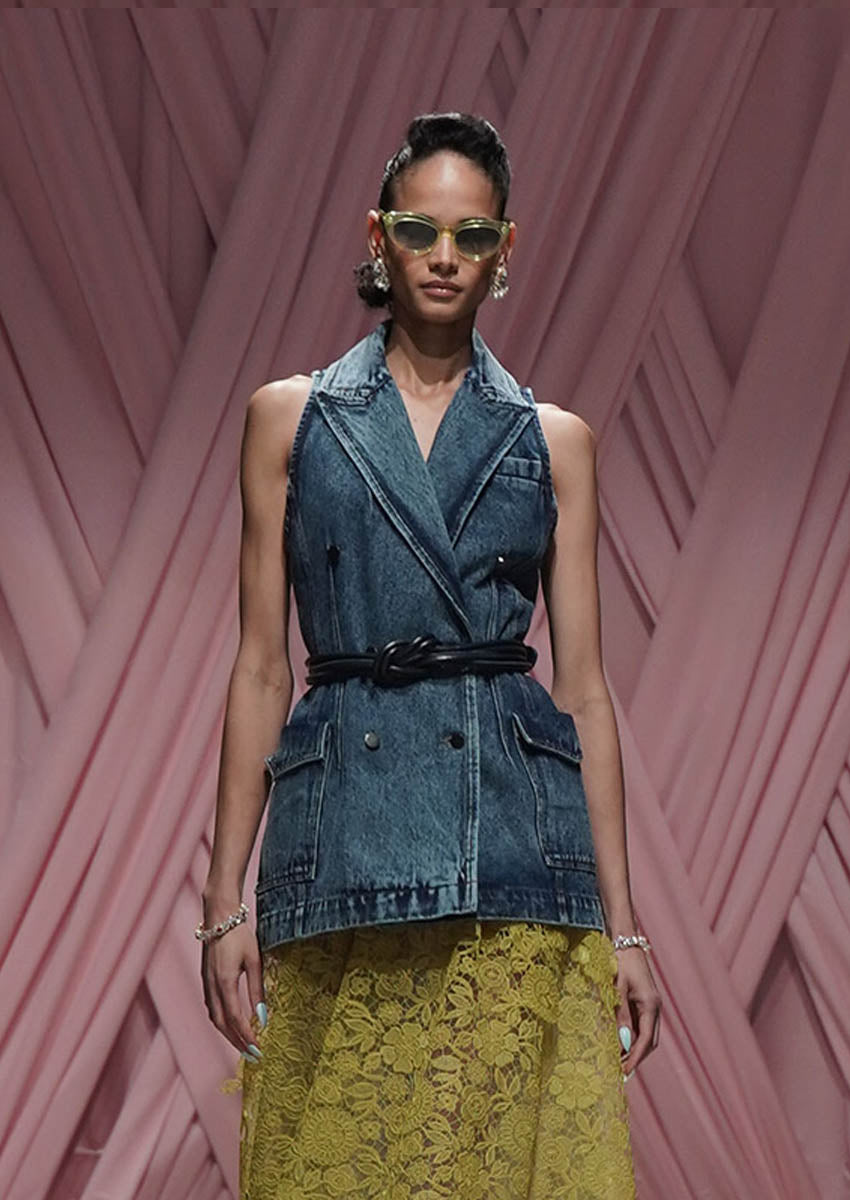 Model wearing a denim vest over a yellow dress against a pink geometric background