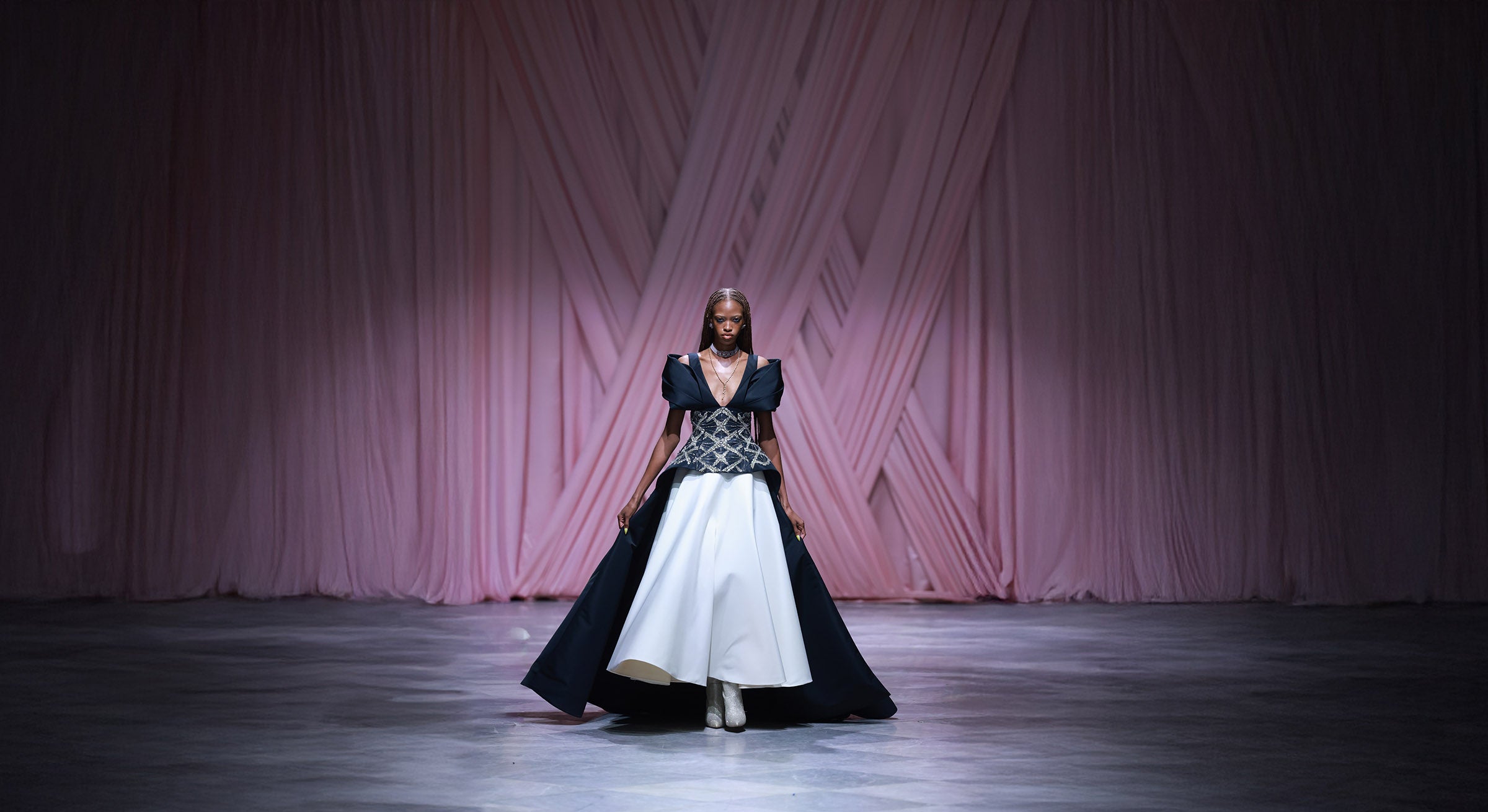Model wearing a black and white dress on a runway with a pink geometric patterned curtain in the background.