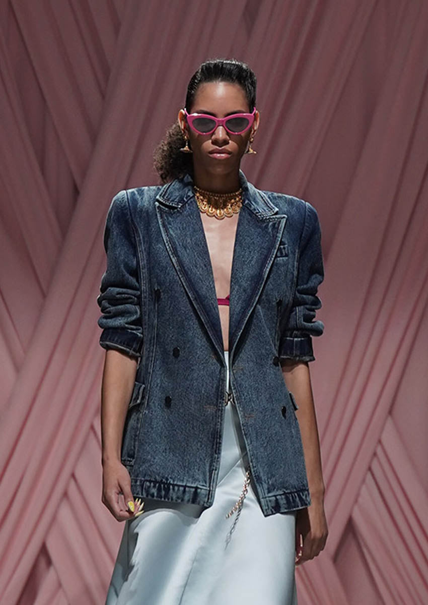 Model wearing a denim jacket and sunglasses against a pink geometric patterned background