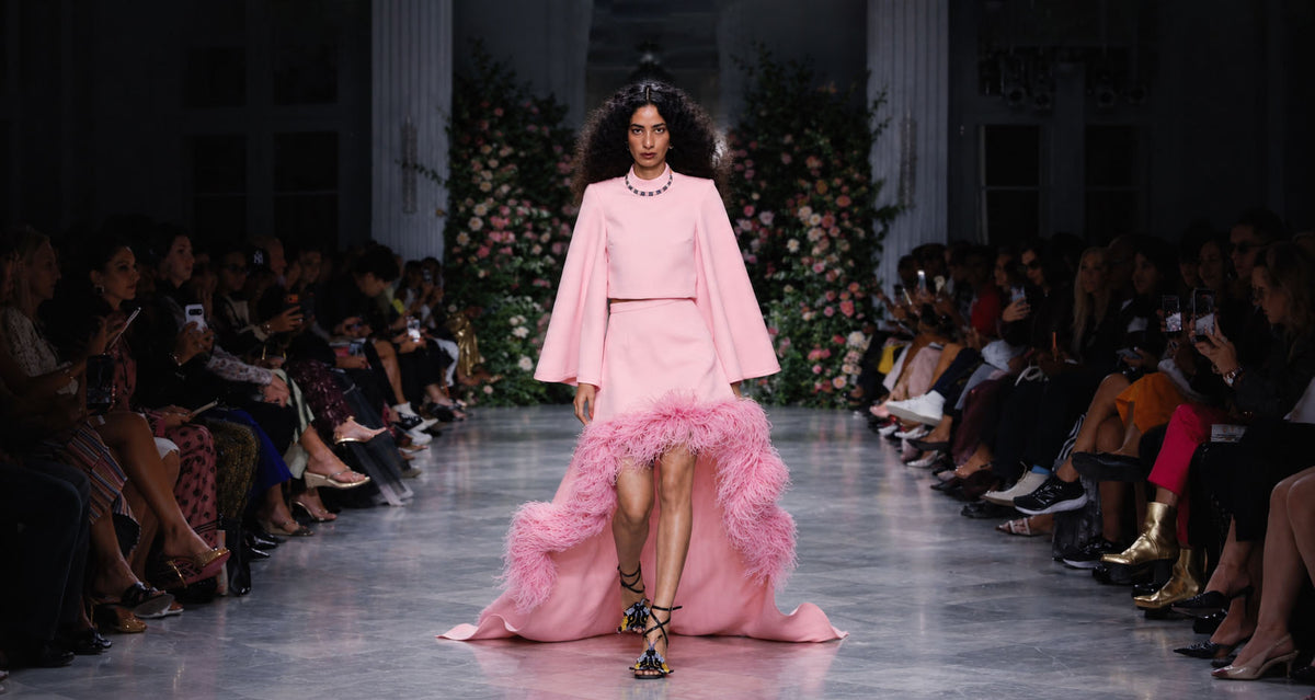 A model walks down a fashion runway wearing a light pink two-piece outfit with dramatic long sleeves, a fitted waist, and a voluminous feathered hem. The runway is lined with seated spectators, and a floral backdrop stands behind the model.